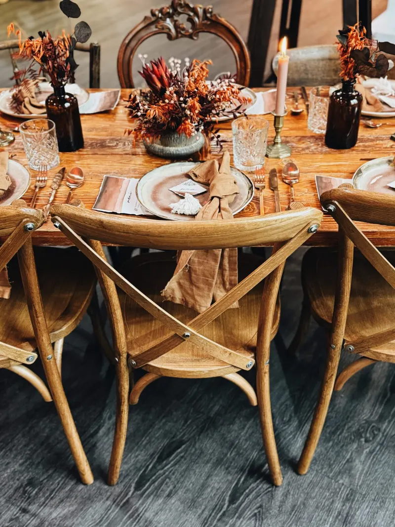 Crossbackchairs aus hellem Holz mit Lederriemen an einer gedeckten Holztafel mit Trockenblumen