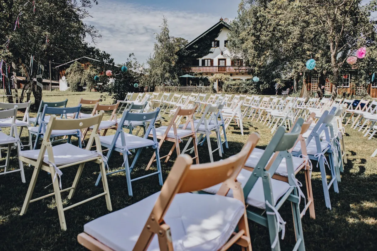 Pastellfarbene Holzklappstühle in Weiß, Blau, Rosa und Natur in Reihen vor einem Landhaus mit Lampions