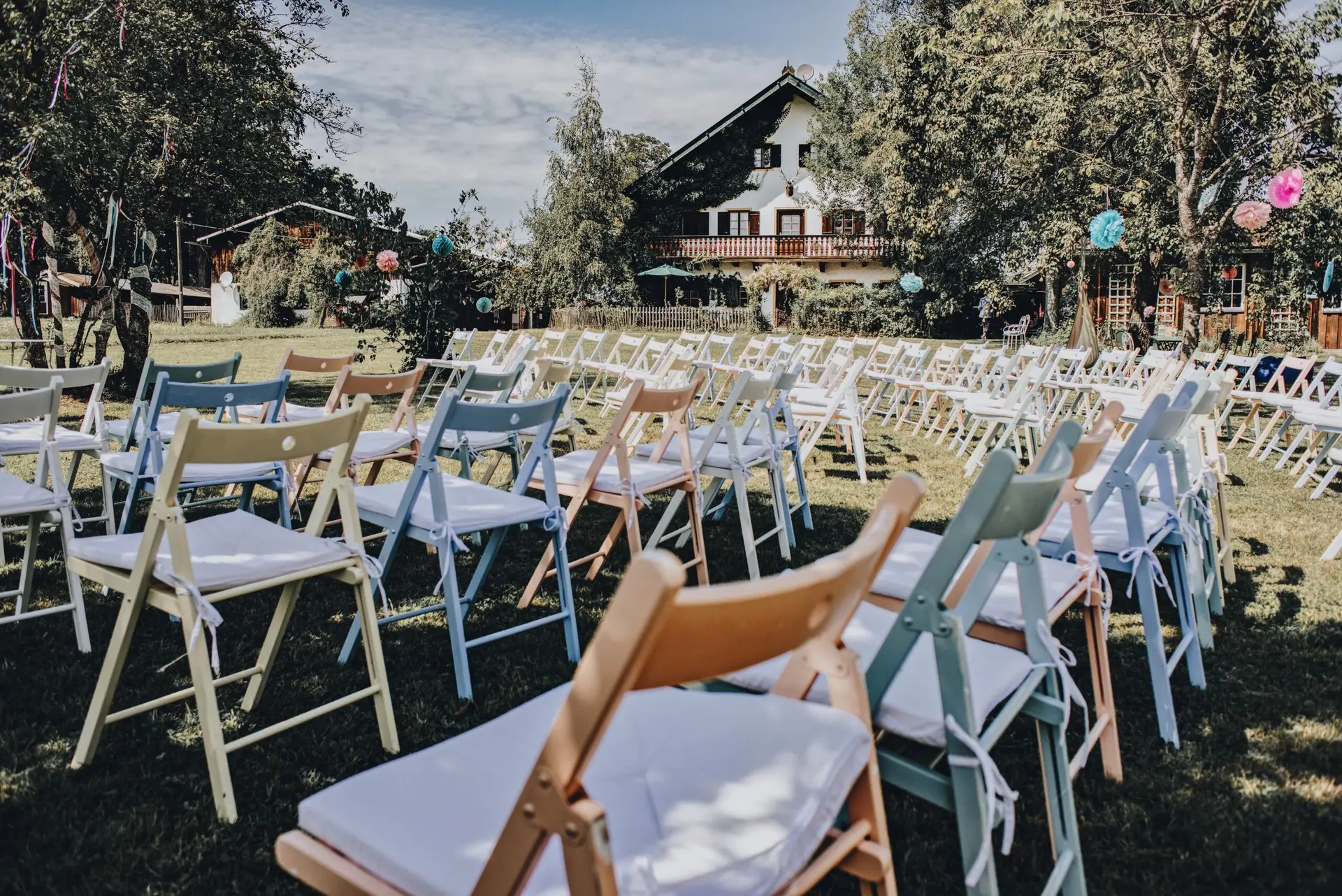 Pastellfarbene Holzklappstühle in Weiß, Blau, Rosa und Natur in Reihen vor einem Landhaus mit Lampions
