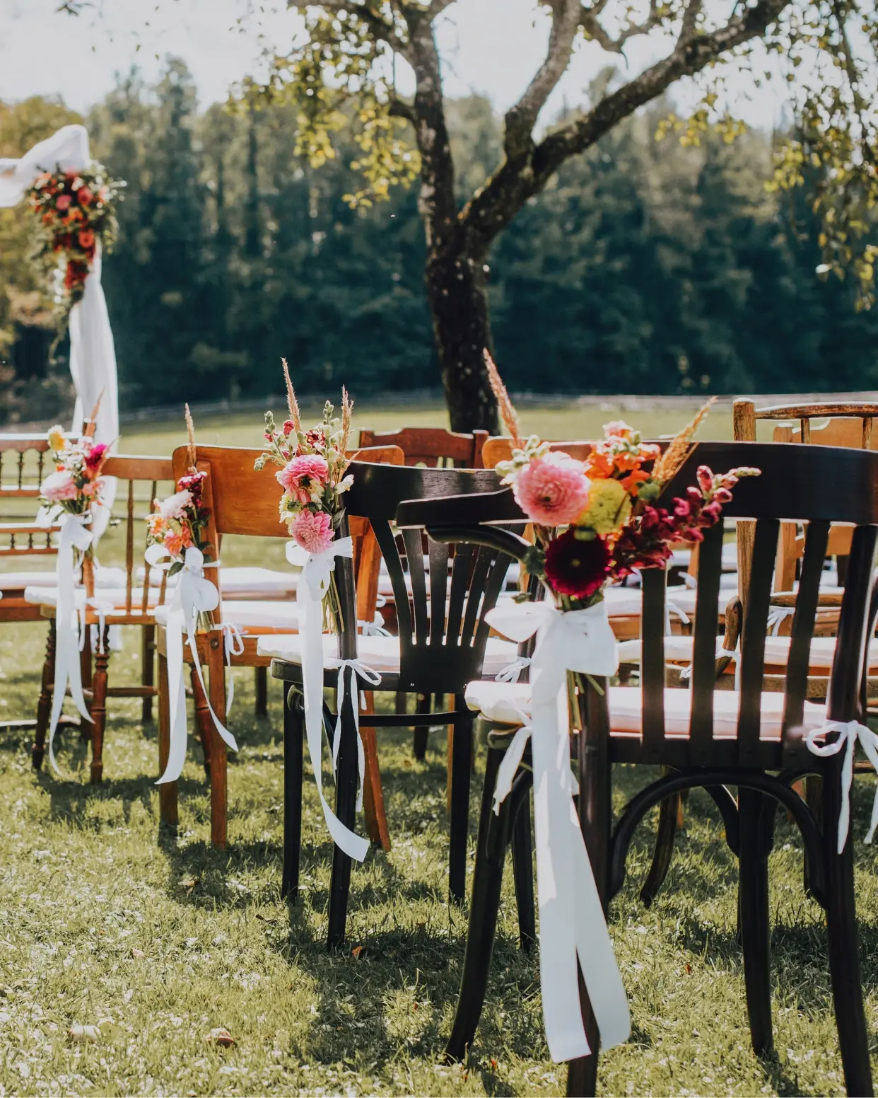 Gemischte Vintage Holzstühle mit Blumenschmuck und weißen Bändern bei einer freien Trauung im Garten